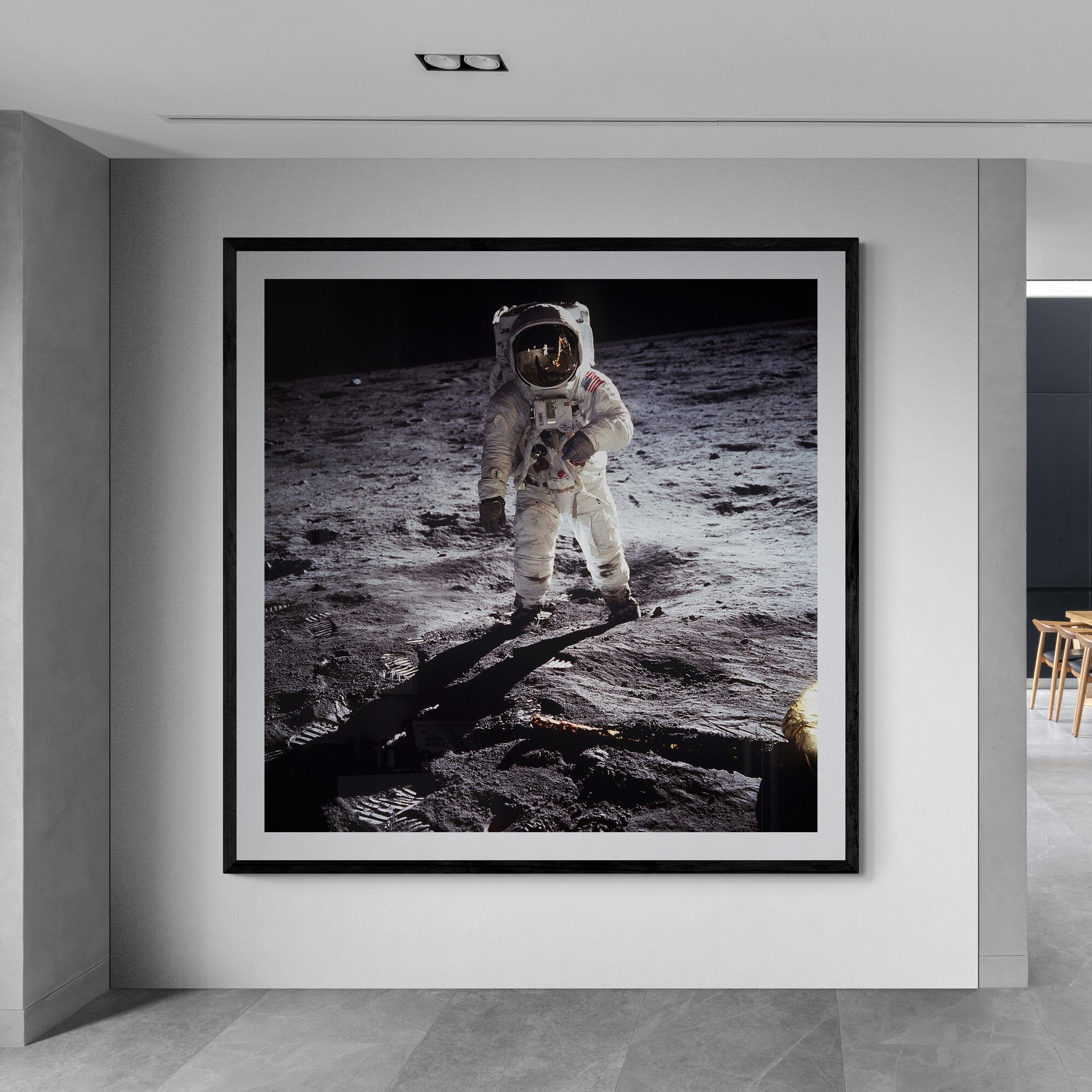 A large framed black and white photograph of an astronaut on the moon, displayed on a light gray wall in a modern interior hallway with a dining area visible in the background.